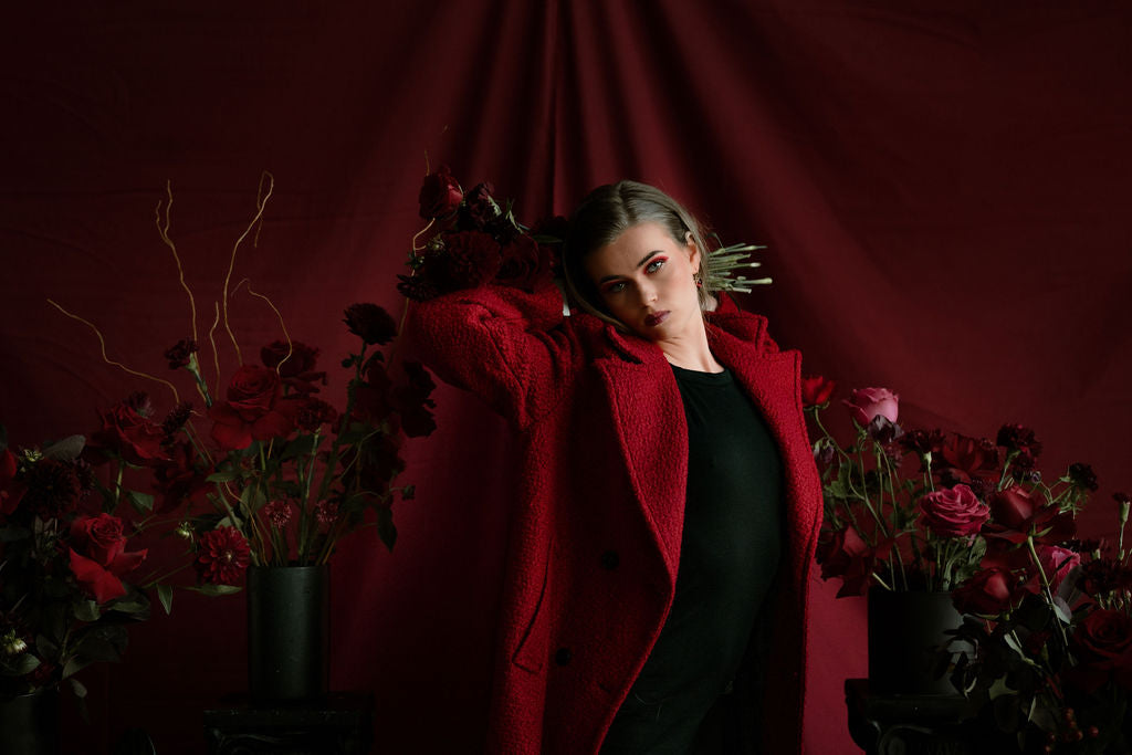 Woman in a red coat standing between red flowers against a dark red curtain. Photo by Lynette Photographer, makeup by Len, and Grace Beauty Salon
